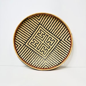 Puede incluir: Una bandeja redonda tejida con un patrón geométrico en blanco y negro. La bandeja tiene un borde marrón.