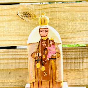 Może przedstawiać: Drewniana figurka kobiety w złotej i brązowej szacie, trzymającej małe dziecko w ramionach. Kobieta ma na głowie koronę i aureolę. Figurka stoi na różowym cokole z napisem "CARMEN".