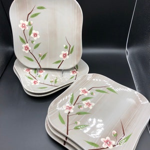 May include: A set of four square ceramic dinner plates with a light gray background and a floral design. The design features a branch with pink flowers and green leaves.