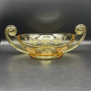 May include: A vintage yellow glass serving bowl with two ornate handles. The bowl features etched floral and geometric designs. The handles have a spiral design at the ends. The bowl is on a dark surface.