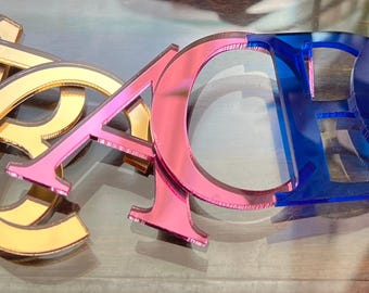 May include: A set of three-dimensional acrylic letters spelling the word BEACH. The letters are finished in reflective metallic gold, vibrant pink, and translucent blue, arranged in a horizontal row with a smooth, glossy surface texture.
