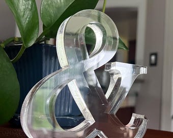 May include: A clear acrylic ampersand symbol, standing upright on a brown surface. The symbol is in front of a blue pot with green leafy plants. The ampersand is a decorative item, suitable for home or office decor.