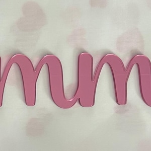 May include: A custom name sign featuring the name Emmy written in a cursive script font. The lettering is crafted from a smooth, reflective pink material with a glossy finish, creating a distinct three-dimensional appearance against the surface.