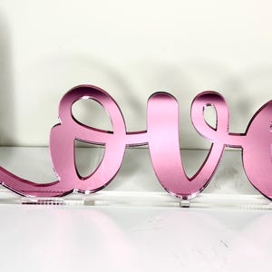 Könnte beinhalten: Ein pinkfarbenes, verspiegeltes "Love"-Schild in Schreibschrift. Das Wort ist auf einer transparenten Acrylbasis montiert, wodurch ein Schwebeeffekt entsteht. Das Schild wird vor einem weißen Hintergrund angezeigt.