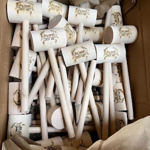 May include: A collection of light-colored wooden mallets packed inside a cardboard box. Each mallet head features a branded logo with a crab illustration and the text JOE WALEGA burned into the wood. The mallets have cylindrical heads and handles.