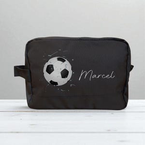 Op de afbeelding: Zwarte rechthoekige toilettas met een voetbalontwerp en de naam "Marcel" in witte letters. De voetbal is wit en zwart met grijze spetters. De tas heeft een handvat aan de zijkant.