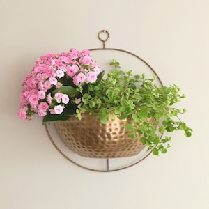 Op de afbeelding: Een gouden metalen hangende plantenpot met een gehamerde textuur bevat roze en groene planten. De pot hangt aan een rond metalen frame.