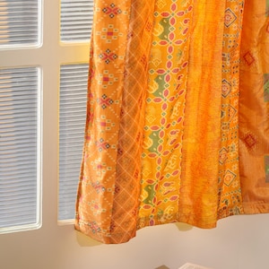 Könnte beinhalten: Orangefarbene Vorhangpaneele mit geometrischen und floralen Mustern hängen in der Nähe eines Fensters. Die Vorhänge sind aus leichtem Stoff gefertigt und verdecken das Fenster teilweise. Ein Buch liegt auf einer gelben Ottomane.