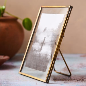 May include: A gold-toned metal picture frame with a glass front and a stand. The frame is holding a black and white photo of a person walking in the rain.