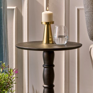 May include: A black wooden side table with a round top and a single pedestal base. A gold candle holder with a white candle sits on the table, next to a clear glass filled with water.