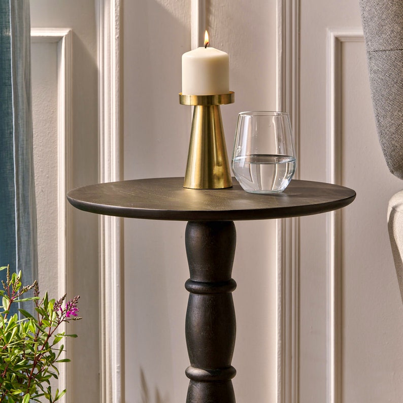 May include: A black wooden side table with a round top and a single pedestal base. A gold candle holder with a white candle sits on the table, next to a clear glass filled with water.