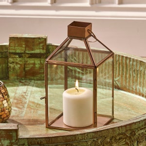 May include: A copper lantern with a glass enclosure and a white pillar candle inside. The lantern is sitting on a wooden tray with a green patina.