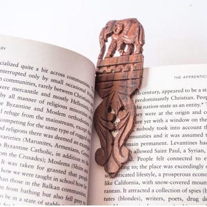 Puede incluir: Un marcapáginas de madera marrón oscuro con un elefante tallado en la parte superior y volutas florales detalladas que se extienden a lo largo de la pieza, colocado entre las páginas de un libro abierto con texto.