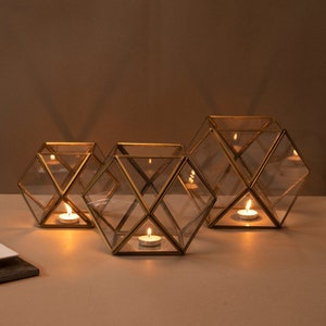 Könnte beinhalten: Drei geometrische Kerzenhalter mit einem goldfarbenen Metallrahmen und transparenten Glasplatten. Jeder Halter enthält eine brennende Teelichtkerze, die ein warmes Leuchten erzeugt. Die Kerzenhalter sind auf einer hellen Oberfläche angeordnet.
