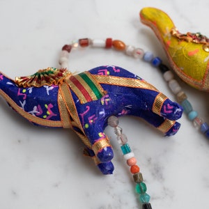 Könnte beinhalten: Eine bunte Perlenkette mit drei kleinen Stoffelefanten. Die Elefanten sind mit goldenen Verzierungen verziert und haben eine blaue, gelbe und grüne Farbgebung.