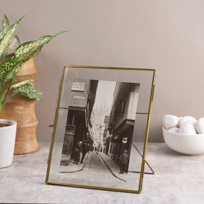 May include: A gold-toned picture frame with a black and white photo of a street scene. The photo shows a narrow street with buildings on either side. The text "MAY DOVILES IN & OUT LIMITED YOUR LAW" is visible on a sign in the photo.