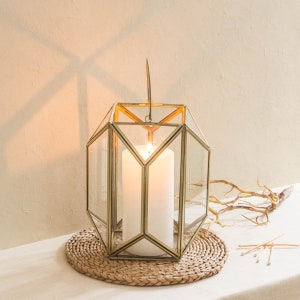 May include: A gold geometric candle lantern with a clear glass body and a white candle inside. The lantern is sitting on a woven round mat.
