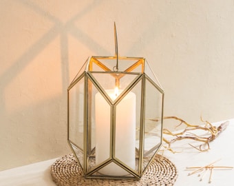 May include: A gold geometric candle lantern with a clear glass body and a white candle inside. The lantern is sitting on a woven round mat.