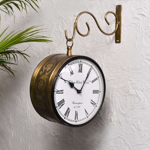 May include: A double-sided wall clock with a brass finish and black accents. The clock face is white with black Roman numerals and hands. The clock is suspended from a decorative brass bracket. The text "Village Clock Works" and "Birmingham est. 1792" are visible.
