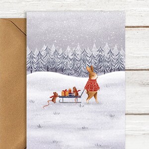 May include: A Christmas card featuring a rabbit in a red sweater pulling a sled with gifts, followed by a mouse. The background shows a snowy forest and falling snow. The card is paired with a brown envelope.