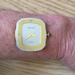 May include: A gold and silver Movado watch with a square face and diamond accents. The watch face is white with black hands and the words "MOVADO" and "QUARTZ" printed in black. The watch also says "SWISS MADE" on the face.