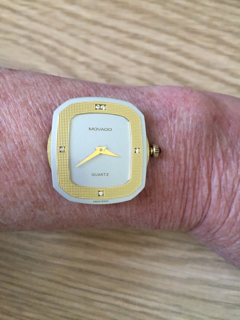 May include: A gold and silver Movado watch with a square face and diamond accents. The watch face is white with black hands and the words "MOVADO" and "QUARTZ" printed in black. The watch also says "SWISS MADE" on the face.
