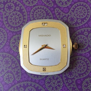 May include: A gold and silver Movado quartz watch with a square face and diamond accents. The watch face is white with black hands and the words "MOVADO" and "QUARTZ" printed in black. The watch also has the words "SWISS MADE" printed in black.