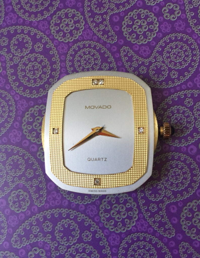 May include: A gold and silver Movado quartz watch with a square face and diamond accents. The watch face is white with black hands and the words "MOVADO" and "QUARTZ" printed in black. The watch also has the words "SWISS MADE" printed in black.