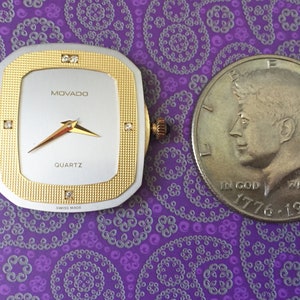 May include: A gold and silver Movado quartz watch face with diamond accents. The watch face is square and has a white background with black hands. The watch face is next to a silver US half dollar coin with the profile of John F. Kennedy.