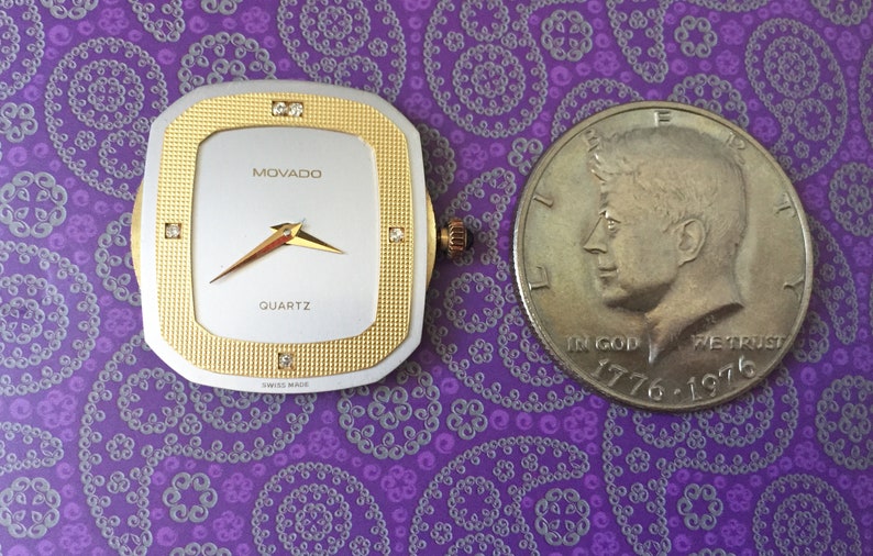 May include: A gold and silver Movado quartz watch face with diamond accents. The watch face is square and has a white background with black hands. The watch face is next to a silver US half dollar coin with the profile of John F. Kennedy.