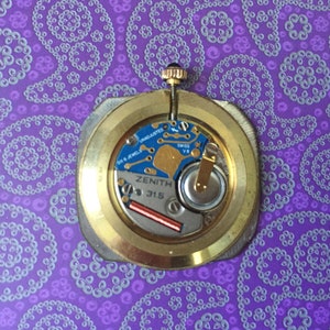May include: Close-up of the back of a gold-toned watch with a Zenith 31.5 movement. The watch has a silver-colored dial with black markings and the text "Zenith 31.5" printed on it.