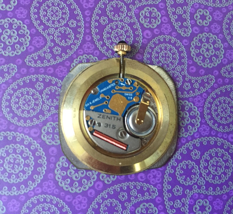May include: Close-up of the back of a gold-toned watch with a Zenith 31.5 movement. The watch has a silver-colored dial with black markings and the text "Zenith 31.5" printed on it.