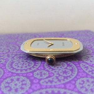 May include: A silver and gold watch with a white face and black hands. The watch has a square shape with rounded corners and a textured bezel. The watch is on a purple patterned surface.