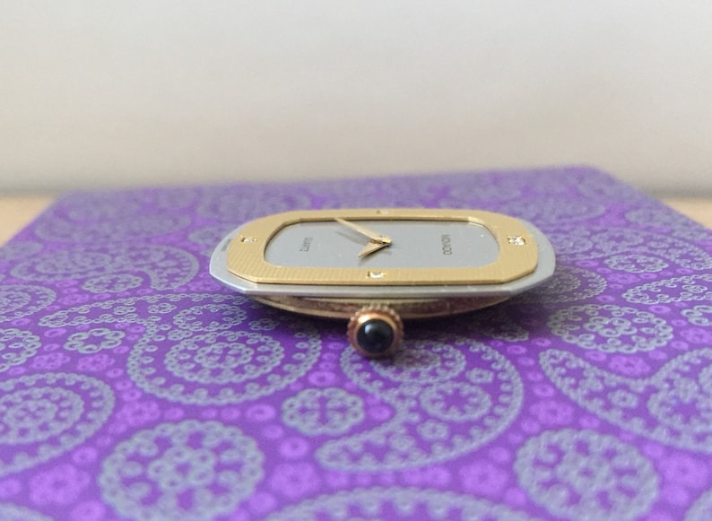 May include: A silver and gold watch with a white face and black hands. The watch has a square shape with rounded corners and a textured bezel. The watch is on a purple patterned surface.