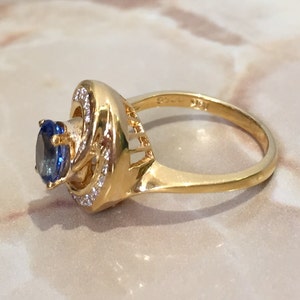 May include: A gold ring with a blue gemstone surrounded by smaller clear gemstones. The ring has a twisted band.