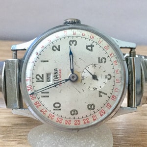 May include: A silver wristwatch with a white face and black hands. The watch has a calendar feature with the day of the week and date displayed. The watch face also has a small sub-dial for the seconds. The watch is on a silver metal band.