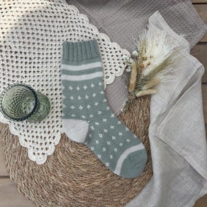 May include: A sage green knitted sock with white accents and a pattern of small white diamonds. The sock features a white heel and toe, and two white stripes near the top. The sock is displayed on a woven mat with a decorative crochet doily.