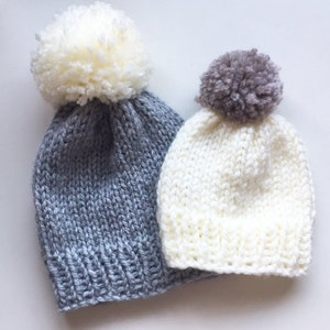 May include: Two knitted hats with pom-poms. The larger hat is gray with a white pom-pom. The smaller hat is white with a gray pom-pom.