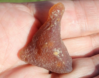 May include: A small, amber-colored, triangular-shaped stone with a textured surface. The stone has a rounded top and is held in an open hand against a backdrop of green grass.