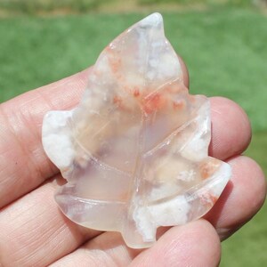 May include: A maple leaf-shaped stone carving. The translucent stone features white, tan, and red hues. The leaf's veins are subtly etched into the surface, creating a naturalistic design. The carving is held in a hand.
