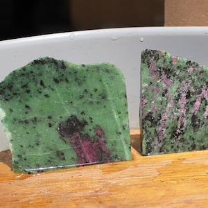 May include: Four rough-cut green, black, and pink gemstone slabs. The slabs are arranged in a row on a wooden surface.