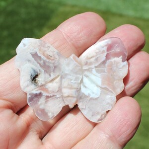 May include: A carved butterfly figurine made of a light-colored stone with pink, white, and brown patterns. The butterfly has detailed wing and body carvings. The figurine is held in a person's hand.