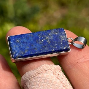 May include: A rectangular lapis lazuli pendant set in a silver-toned metal frame. The stone is a deep blue color with visible gold-colored pyrite flecks throughout the surface. A small silver-toned metal bail is attached to the side of the pendant.