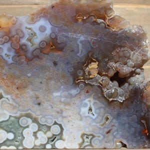 May include: A polished, rectangular agate stone with a complex pattern of colors. The stone features shades of brown, white, gray, and orange, with a bubbly, layered texture. The edges are rough and uneven, showcasing the natural formation.