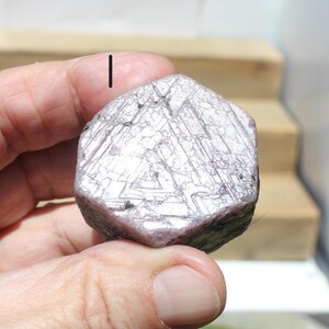 May include: A faceted, light purple gemstone with a complex, geometric pattern. The stone is held in a hand, showcasing its crystalline structure and natural color. The gem's surface reflects light, highlighting its unique texture.