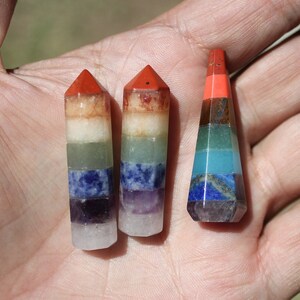 May include: Three small, pointed, multi-colored crystal wands. Each wand is layered with seven different colored stones, representing the seven chakras. The wands are arranged in a row on a hand.