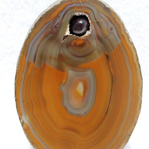 May include: A slice of agate with a deep orange and brown color pattern. The agate has a natural cavity in the center, filled with a dark purple gemstone.