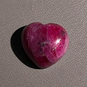 May include: A polished, heart-shaped gemstone in a deep magenta and dark pink hue with visible natural inclusions and a smooth, reflective surface. The stone is positioned near a small, white rectangular marker labeled with the number 1.