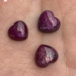 May include: Three small, polished, heart-shaped gemstones in a deep magenta-purple color. Each stone features a smooth, reflective surface with natural internal inclusions. The stones are labeled with the handwritten white numbers 1, 2, and 3.