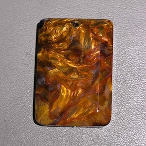 May include: A rectangular tiger's eye gemstone pendant featuring a polished surface with swirling patterns of golden yellow, deep brown, and dark charcoal gray. The stone has a small, circular hole drilled near the top center for suspension.